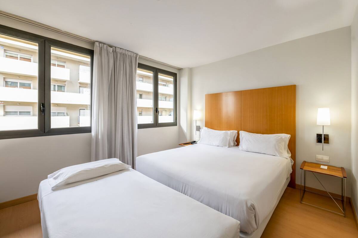 Habitación de hotel con dos camas, cabecera de madera, mesita de noche con lámpara y ventana con cortinas.