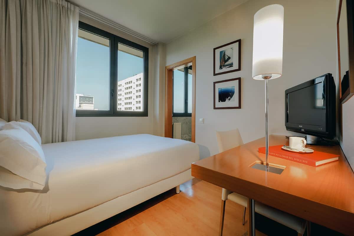 Una habitación de hotel con cama, escritorio, televisión y ventana con cortinas.