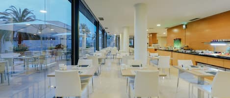 A modern hotel dining area with glass walls, white furniture, and a kitchen visible through the glass.