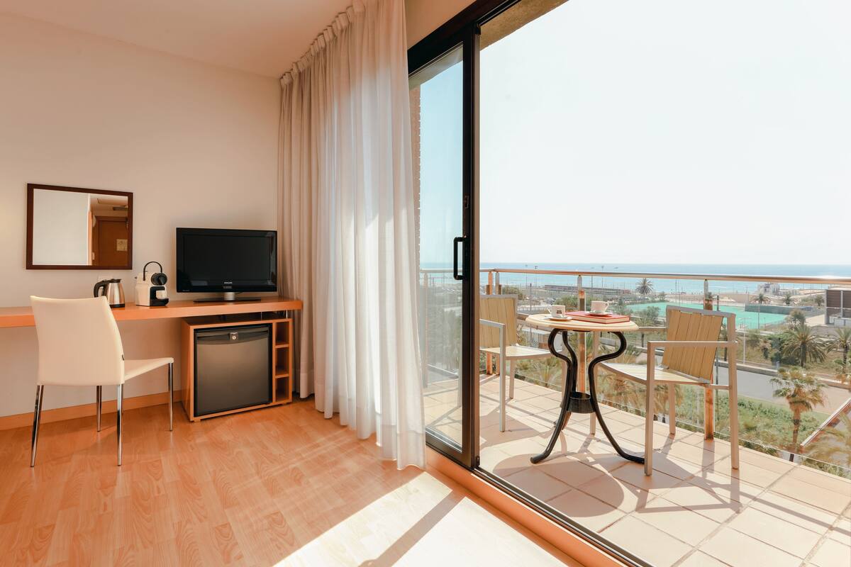 Habitación de hotel con balcón, televisión, escritorio con silla y vistas a la ciudad y la playa.