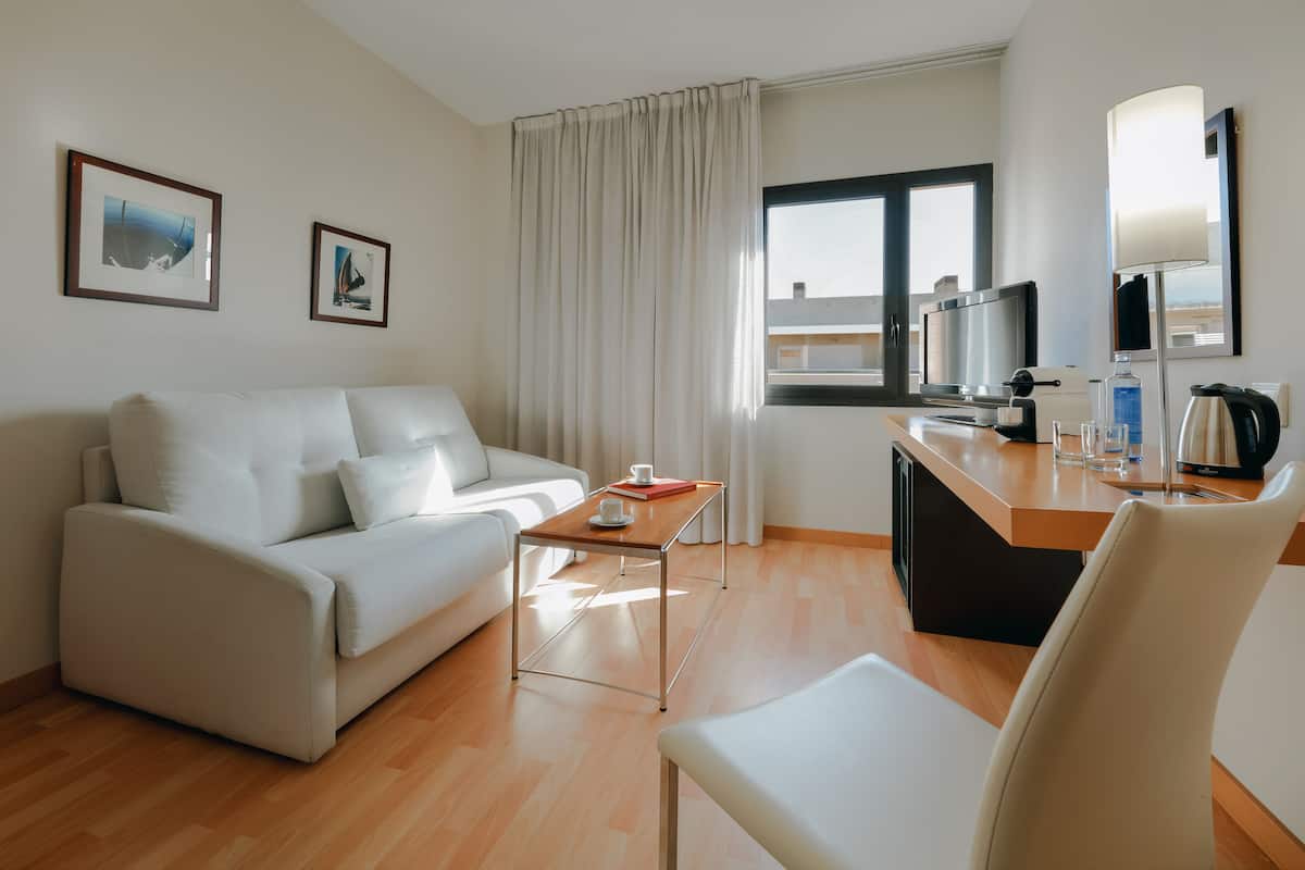 Una habitación de hotel moderna con un sofá blanco, una mesa de centro de madera, un televisor y un escritorio con lámpara y cafetera.
