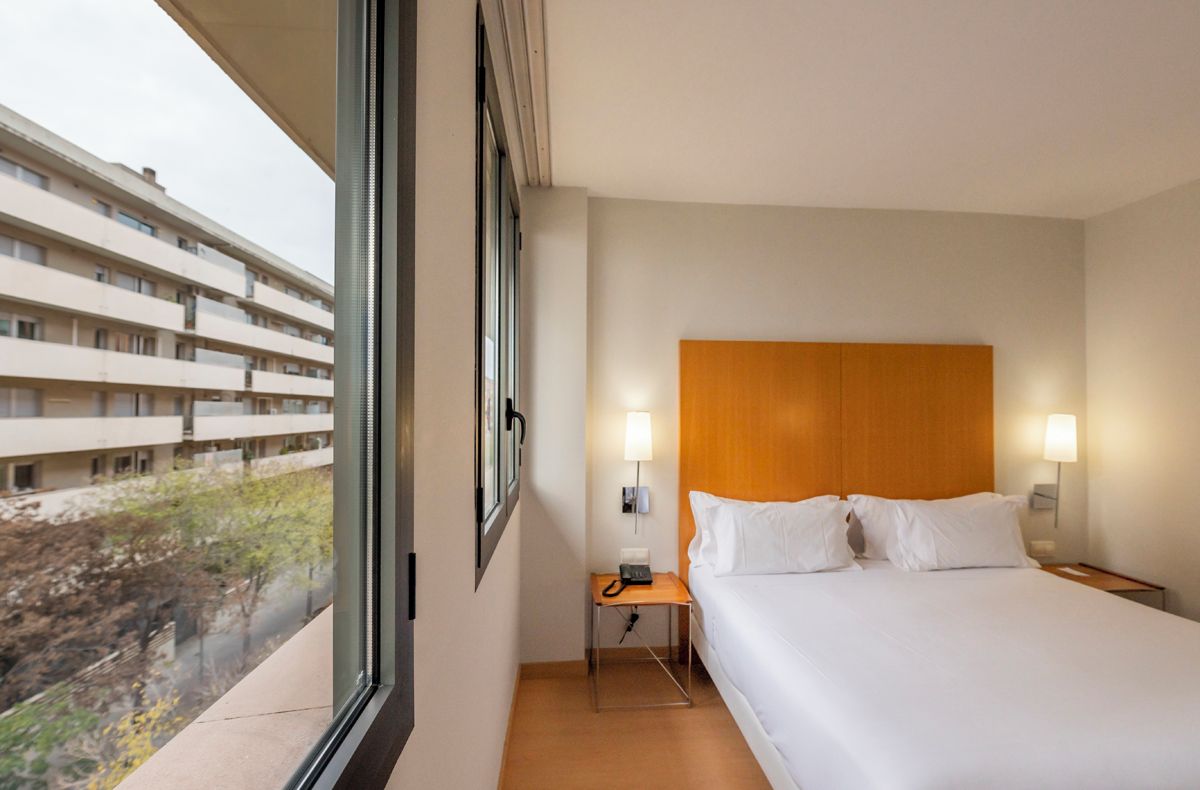 Ein Hotelzimmer mit einem großen Bett, zwei Nachttischlampen, einem Holz-Kopfteil, einem Nachttisch mit Telefon und einem Fenster mit Blick auf ein Gebäude und Bäume.