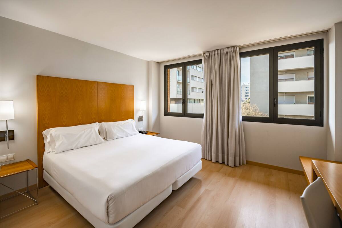 Una habitación de hotel con una cama grande, cabecera de madera, un escritorio, una lámpara y una ventana con cortinas.