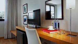 A hotel room with a wooden desk, a flat-screen TV, a mirror, a window with curtains, and a bookshelf with books and bottles.
