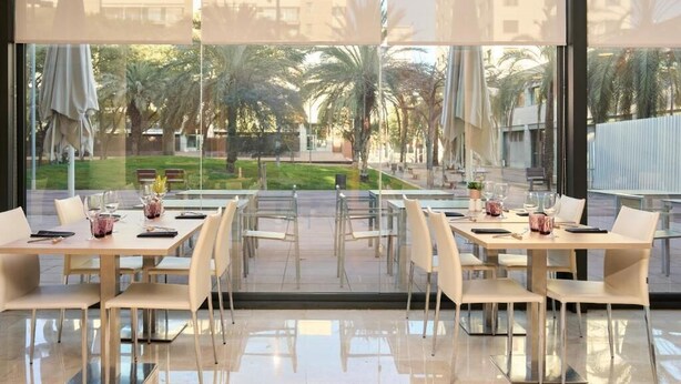 A modern dining area with glass tables and chairs, large windows offering a view of palm trees and a courtyard. - Catalonia Barcelona Beach (Barcelona)