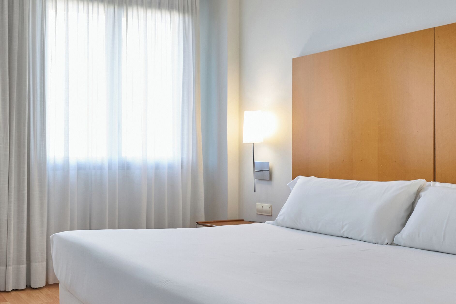 A hotel room with a large bed, white linens, a wooden headboard, a nightstand, a wall-mounted lamp, and sheer curtains.