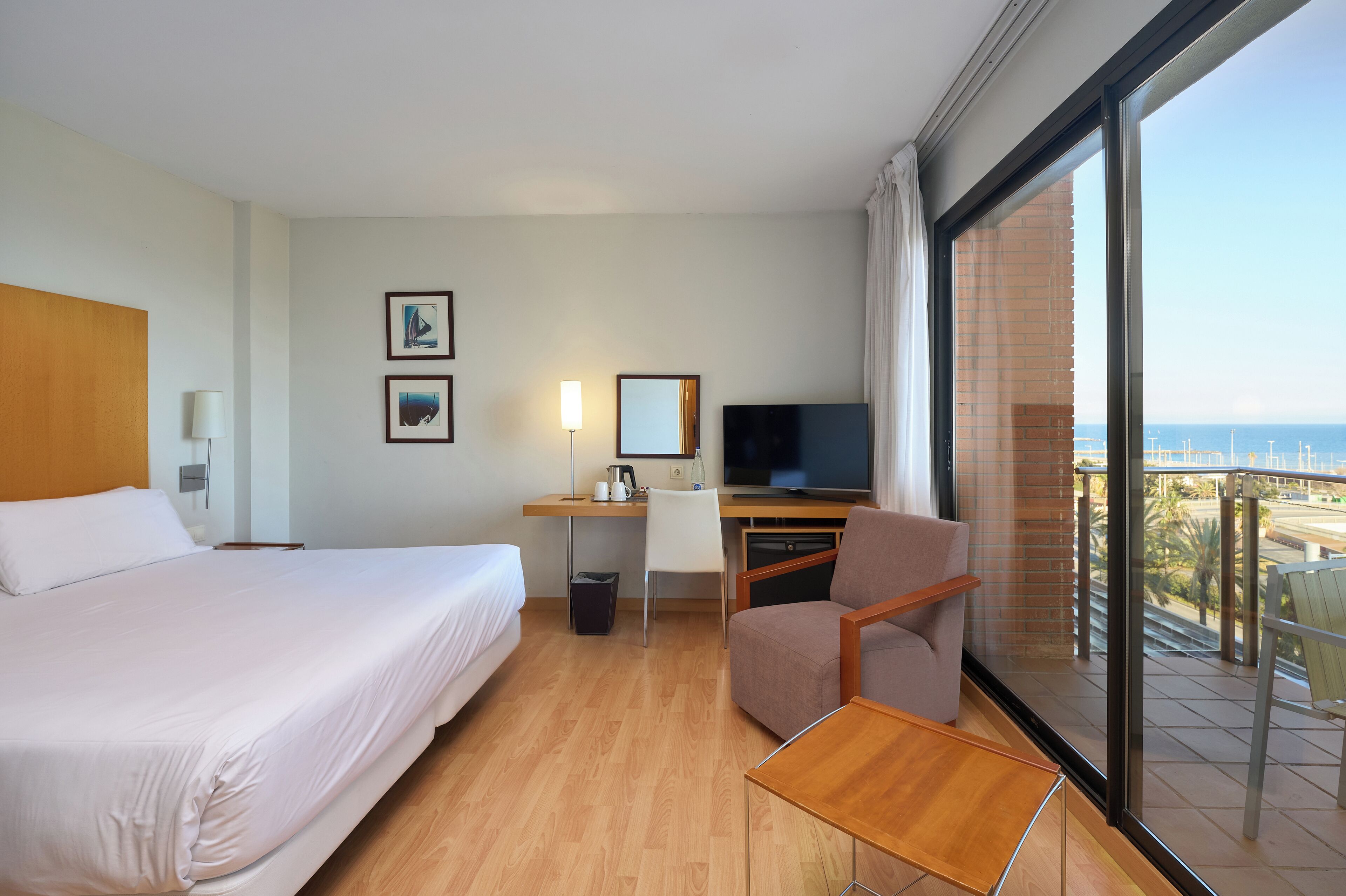 A hotel room with a bed, desk, chair, TV, and a balcony with a view.