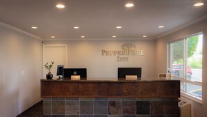 A hotel reception desk with a sign reading "Pepper Tree Inn," a potted plant, and a window with a view of the outside. - PepperTree Inn (Beaverton)