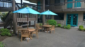 A patio area with wooden tables and chairs, two blue umbrellas, and a stone building with teal accents. - PepperTree Inn (Beaverton)