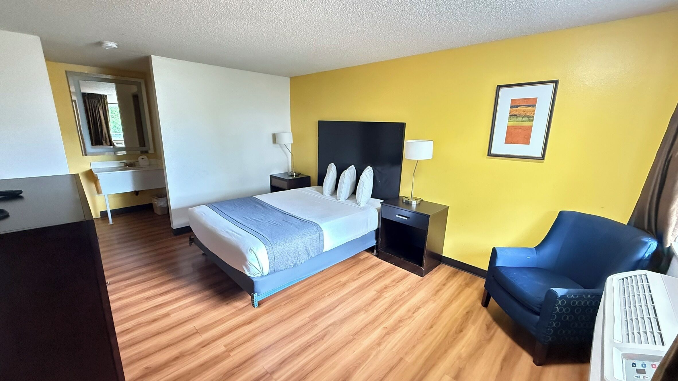 A hotel room with a bed, bedside table, lamp, chair, and a heater.