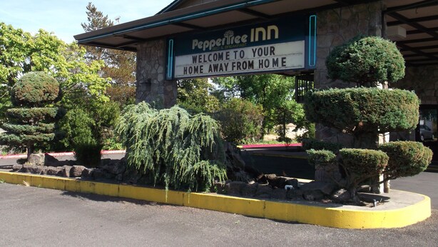 A sign for Peppertree Inn with a welcoming message and a stone building facade. - PepperTree Inn (Beaverton)