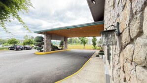 A hotel entrance with a covered parking area, stone facade, and a view of the surrounding landscape. - PepperTree Inn (Beaverton)