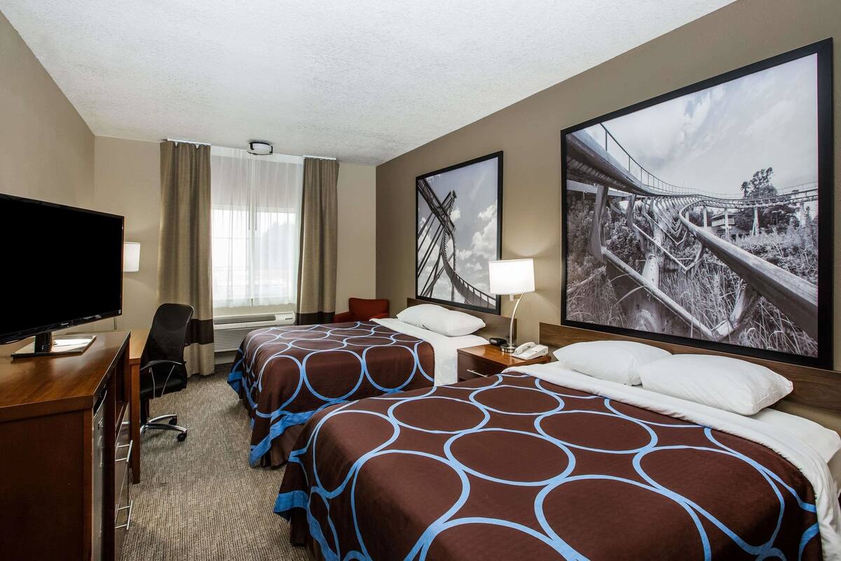 Habitación de hotel con dos camas, un escritorio con televisor, una silla y un ventanal con cortinas.