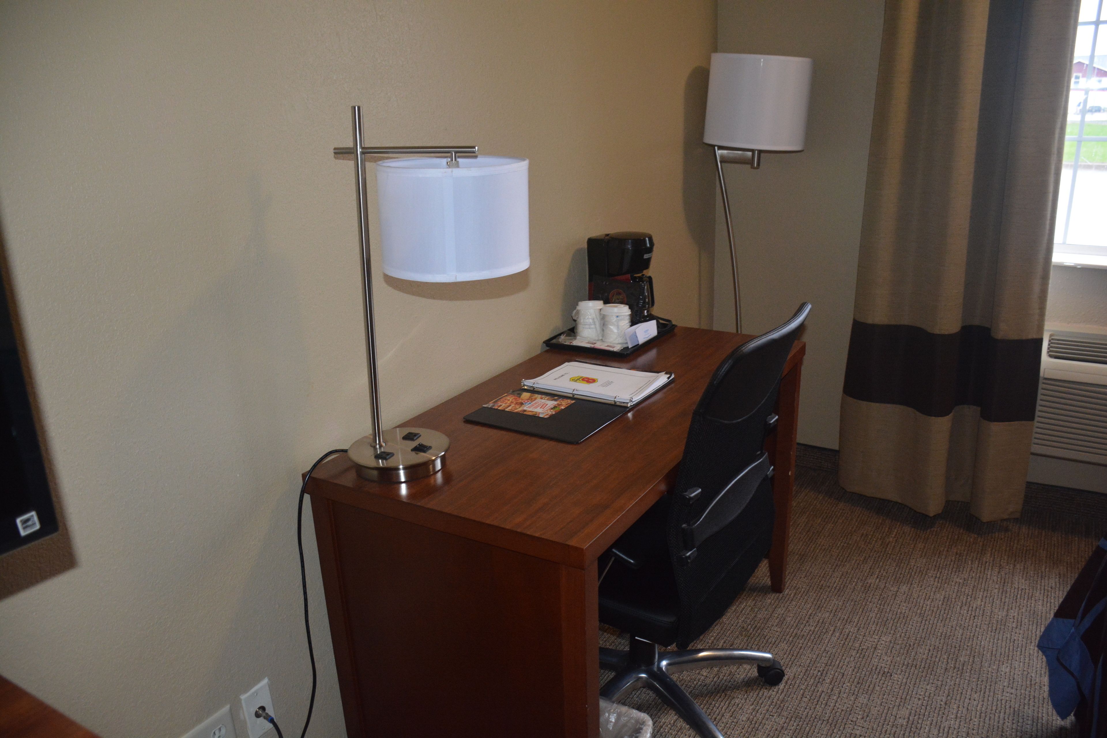 A hotel room with a wooden desk, a lamp, a coffee maker, a chair, and a window with curtains.
