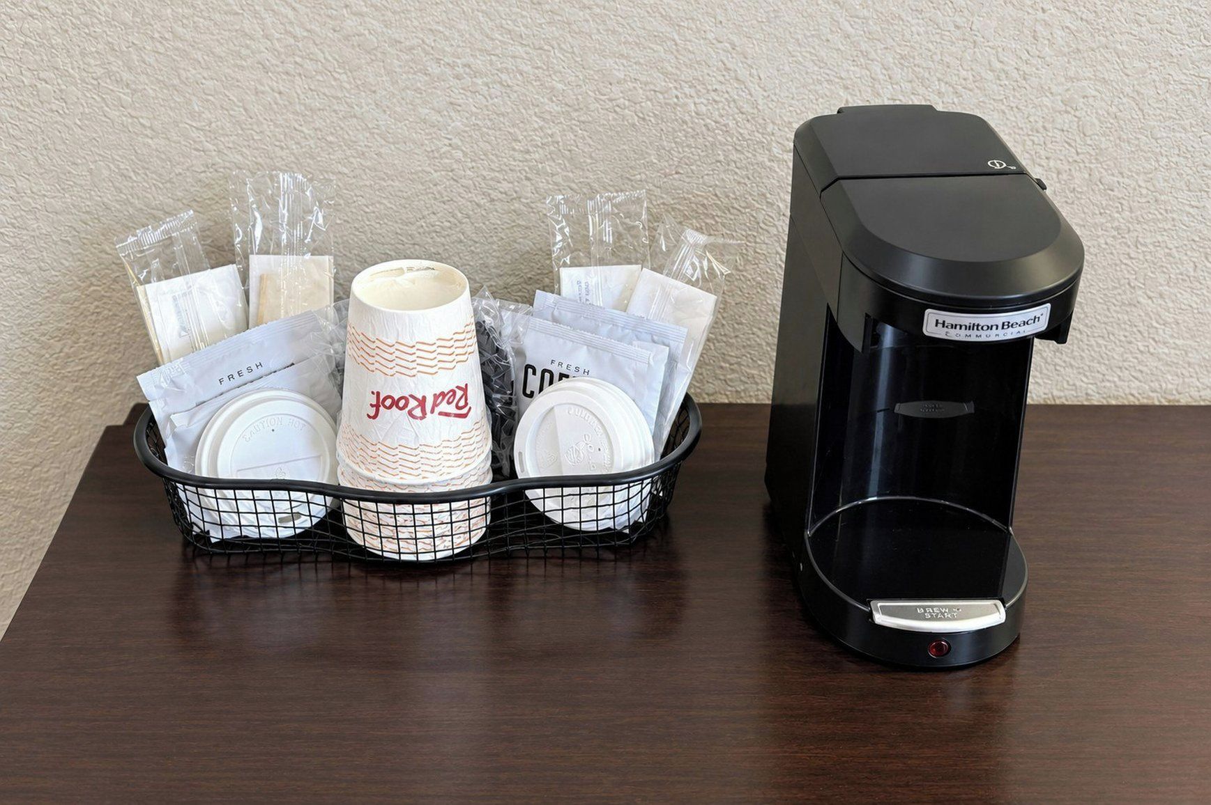 A coffee maker and a basket with disposable cups and napkins on a wooden table.