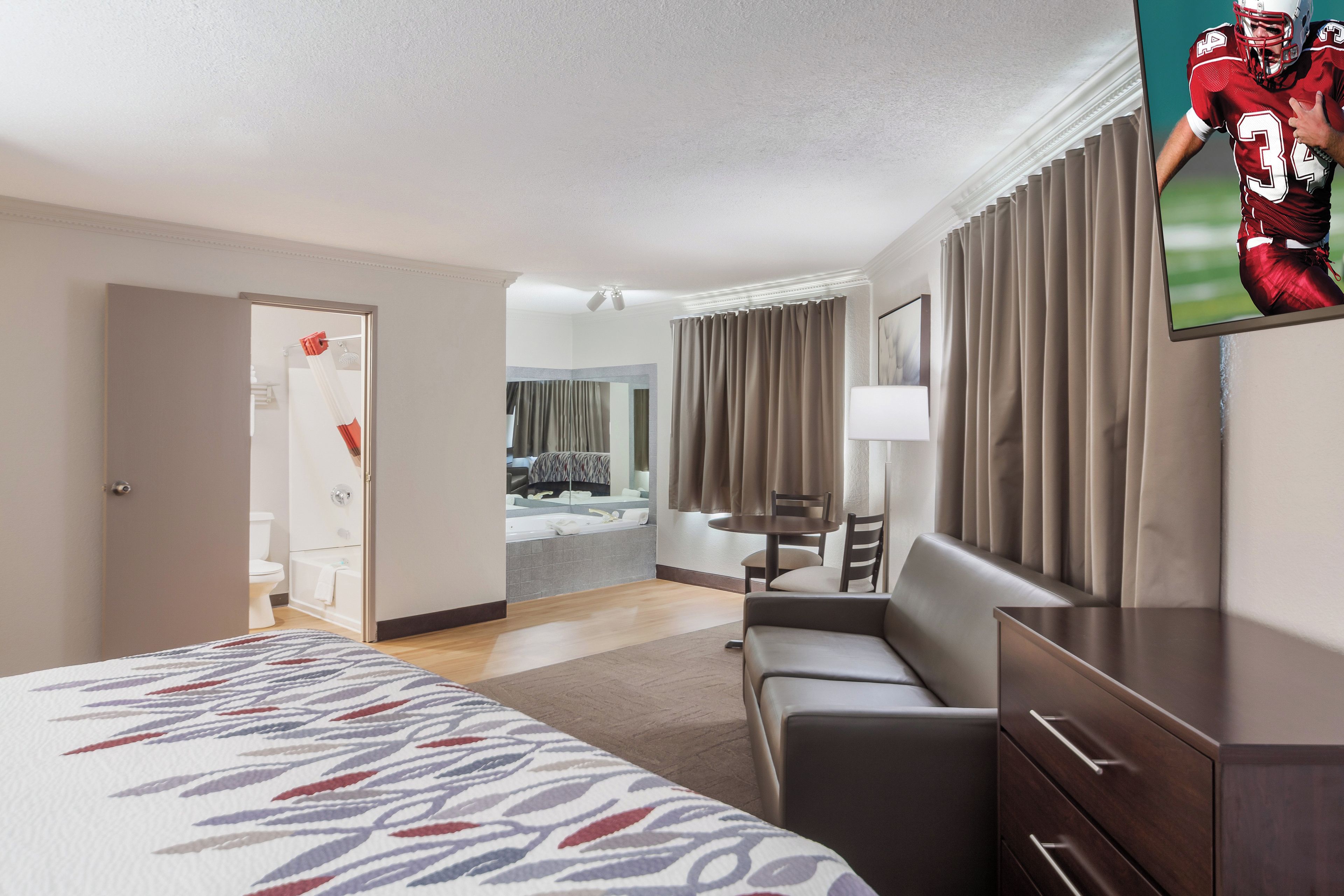 A hotel room with a bed, a desk, a chair, a TV, and a bathroom visible through an open door.