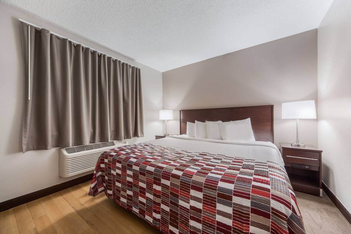 Une chambre d’hôtel avec un grand lit, deux lampes de chevet, une table de chevet, une fenêtre avec des rideaux et un climatiseur fixé au mur.