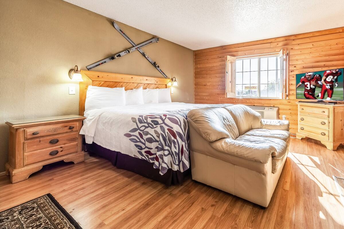 Une chambre avec une tête de lit en bois, un lit avec du linge de maison blanc, un canapé beige, une commode en bois et une fenêtre donnant sur un beau paysage.