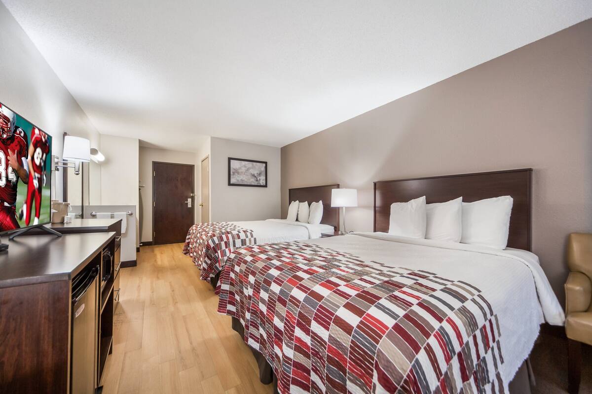 Une chambre d’hôtel avec une kitchenette, deux lits recouverts de couvertures à carreaux rouges et blancs, une tête de lit en bois, une lampe de chevet et un tableau encadré accroché au mur.