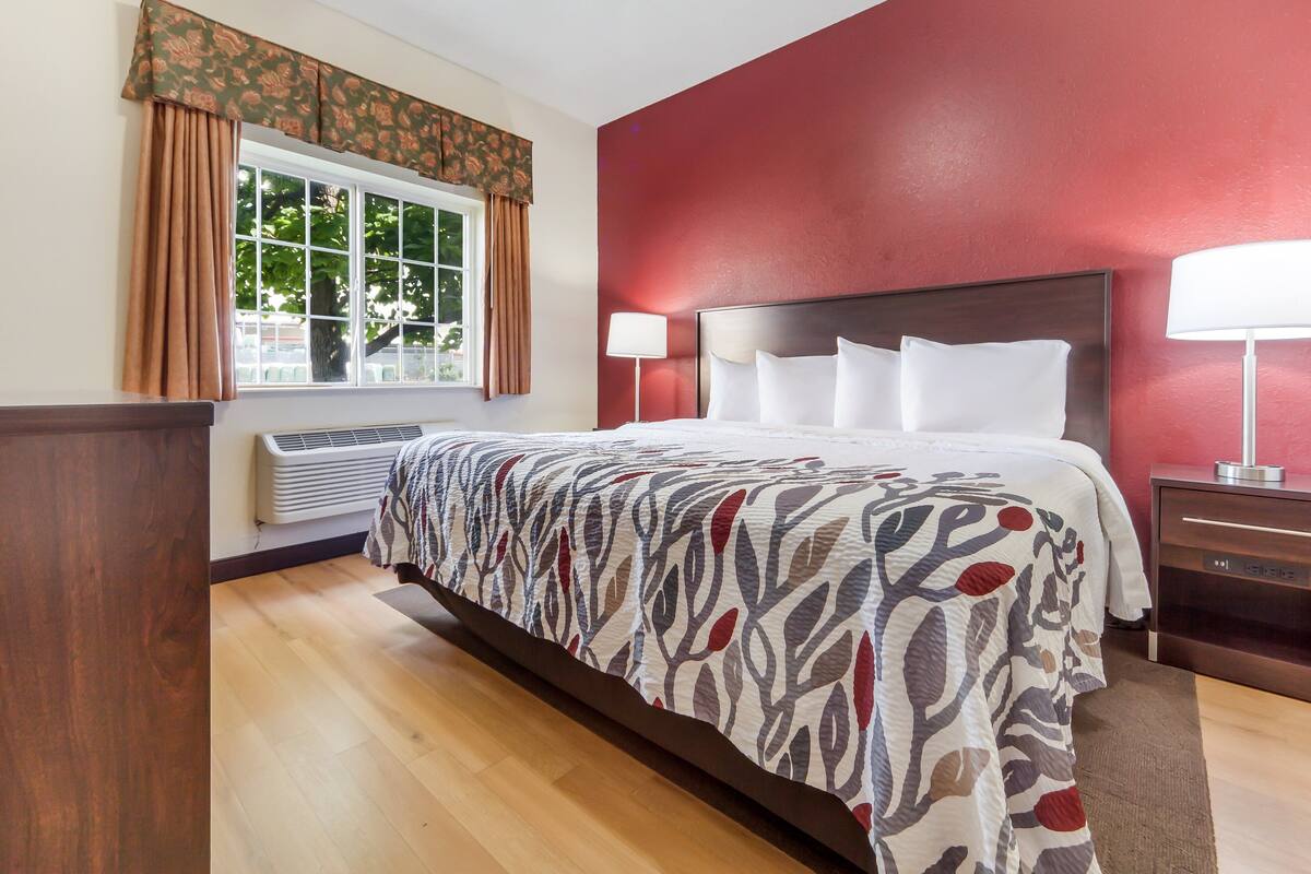 Une chambre d’hôtel avec un grand lit, une fenêtre avec des rideaux, un radiateur et une table de chevet avec une lampe.