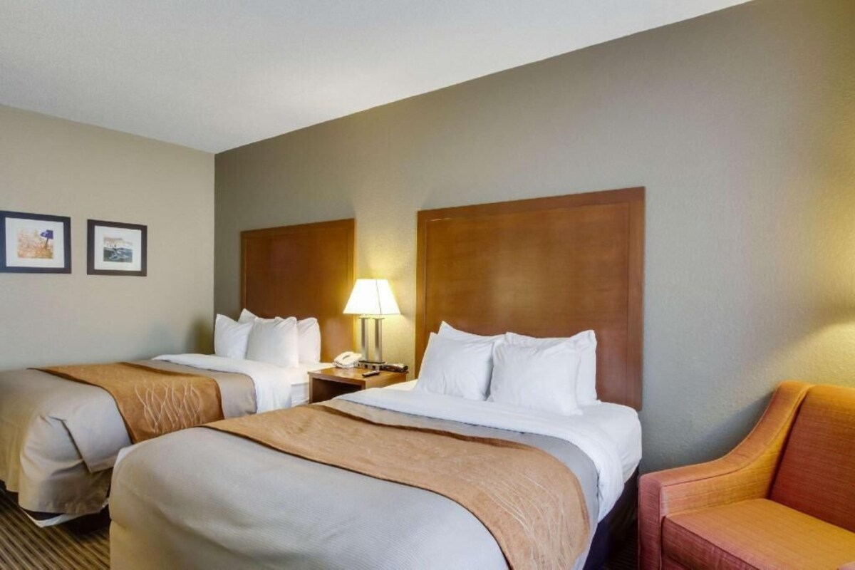 A hotel room with two beds, a nightstand with a lamp, a chair, and framed pictures on the wall.