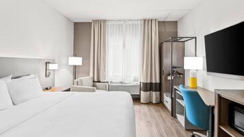Quarto de hotel moderno com uma cama grande, uma mesa com cadeira, uma televisão e uma janela com cortinas.