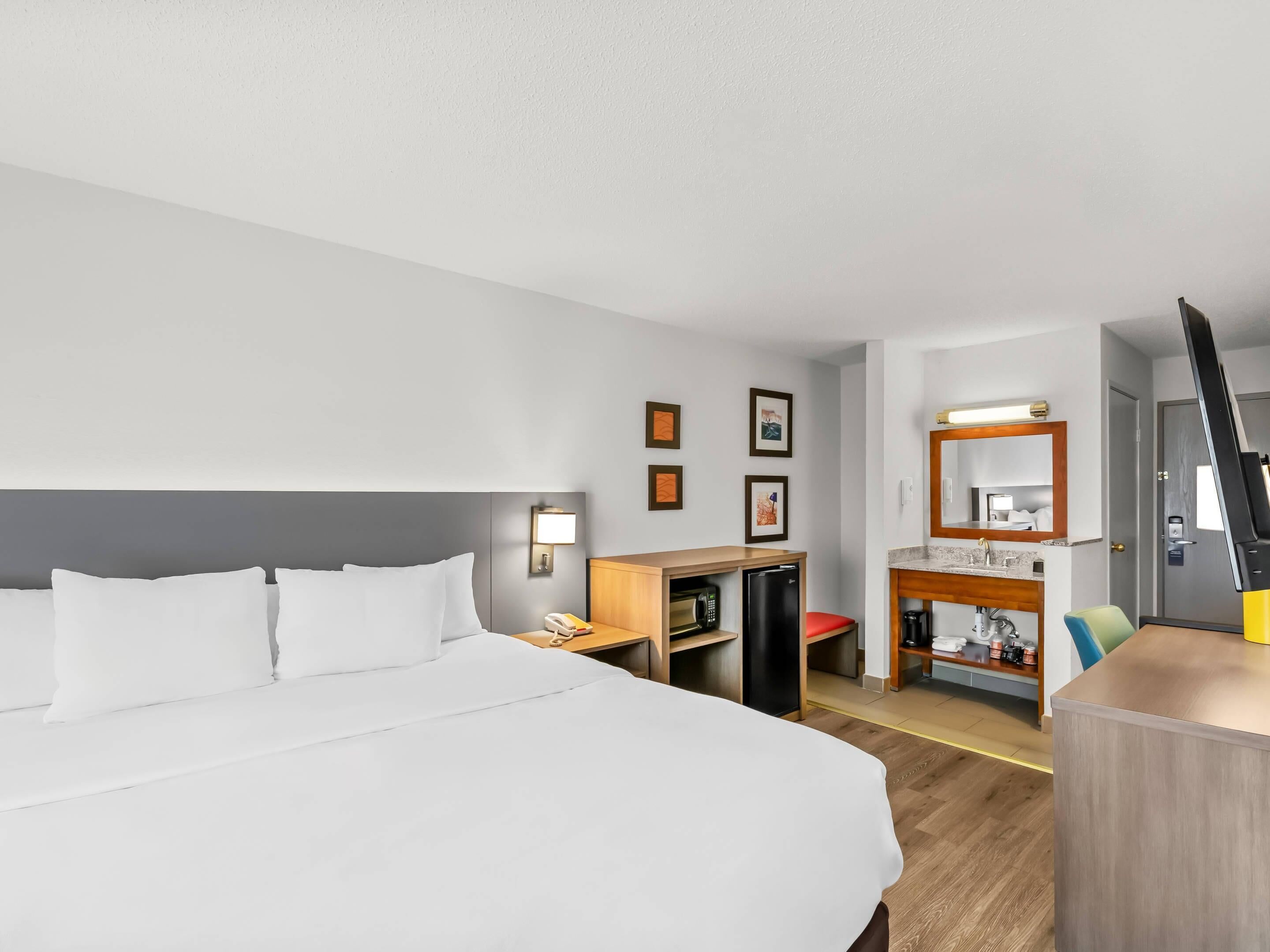 A hotel room with a large bed, a desk with a computer, a small refrigerator, a mirror, and a bathroom with a sink and toilet.