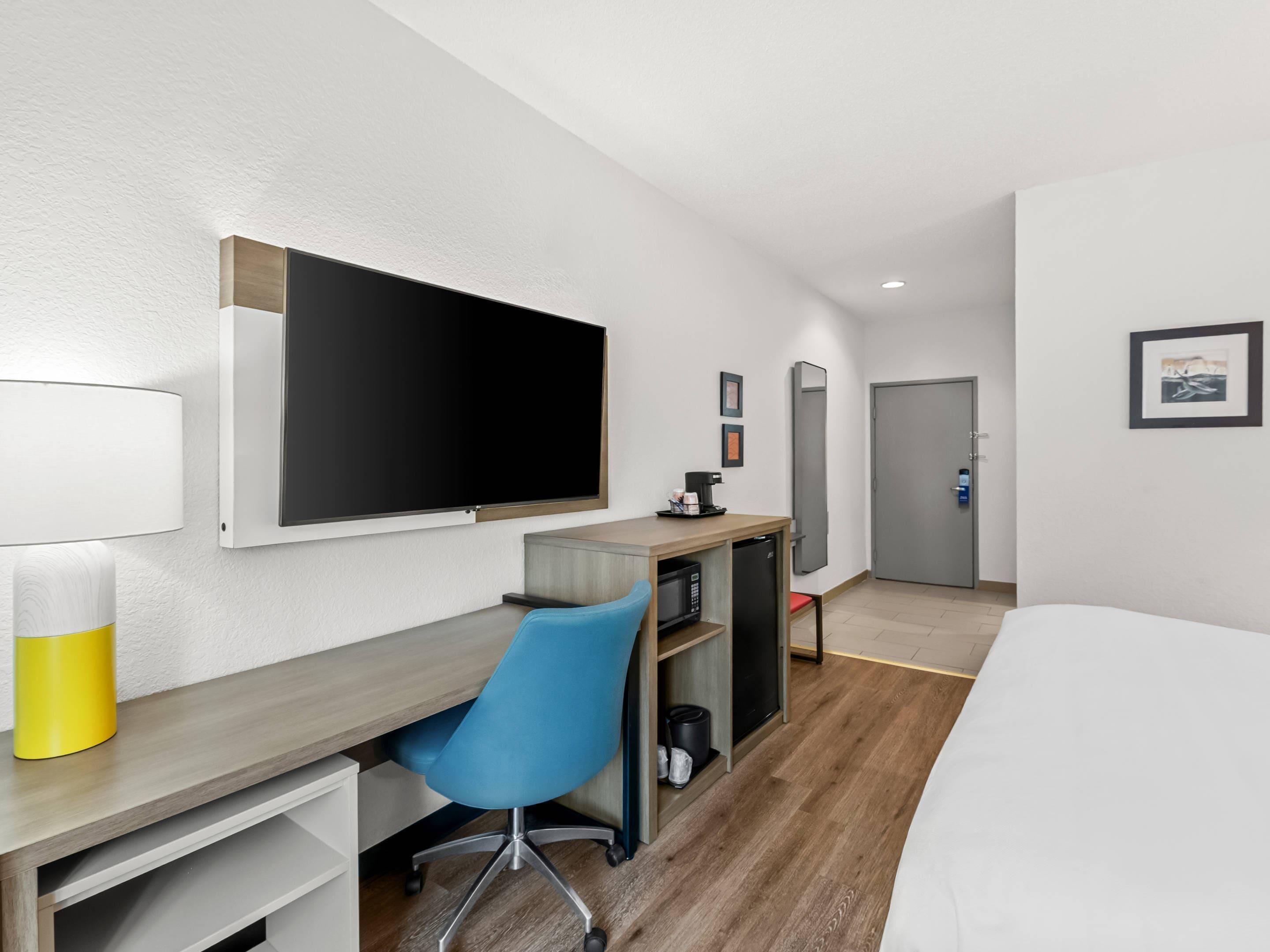 A hotel room with a bed, a desk with a chair, a flat-screen TV, a coffee machine, and a door.