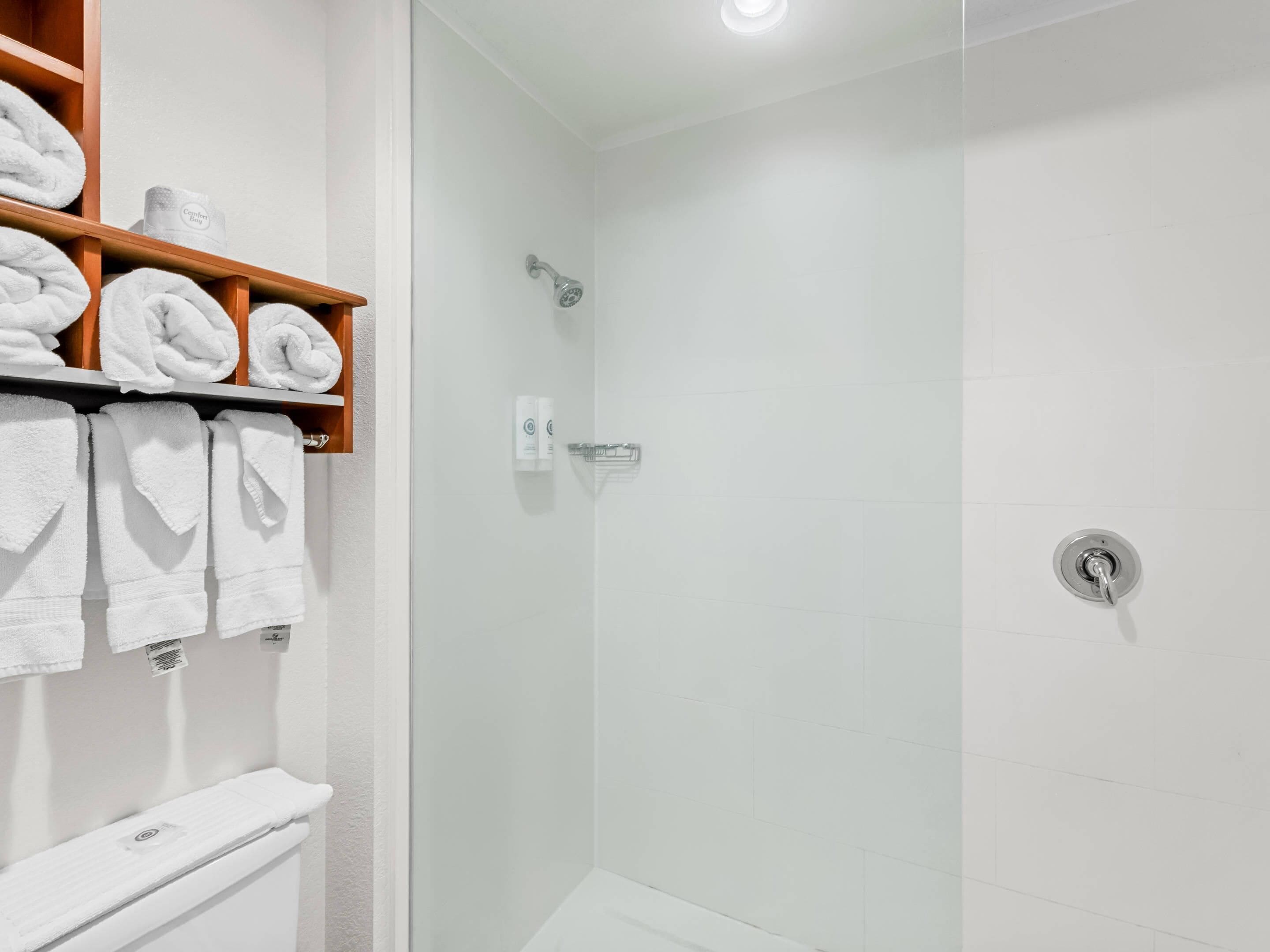 Un baño moderno con cabina de ducha de vidrio, azulejos blancos y un estante de madera con toallas ordenadas.