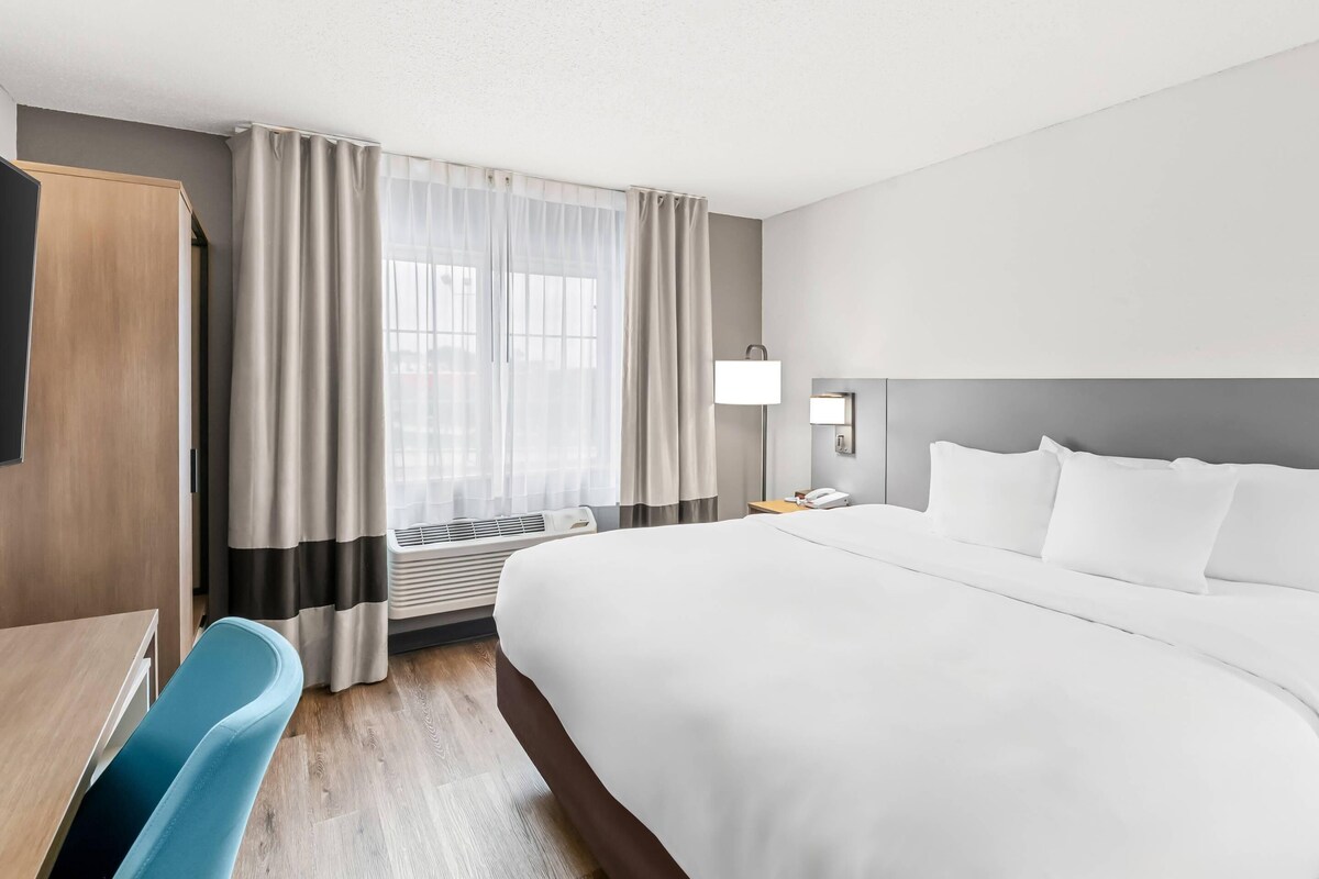 A hotel room with a large bed, a desk, a chair, a desk lamp, and a window with curtains.