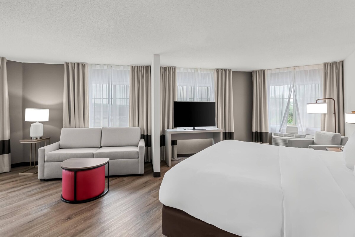 A hotel room with a bed, a sofa, a TV, a desk, and a red side table.