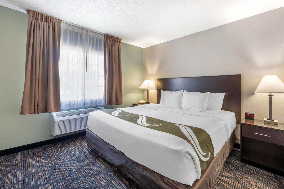 A hotel room with a large bed, two bedside tables, a nightstand, a lamp, a clock, and a window with curtains.