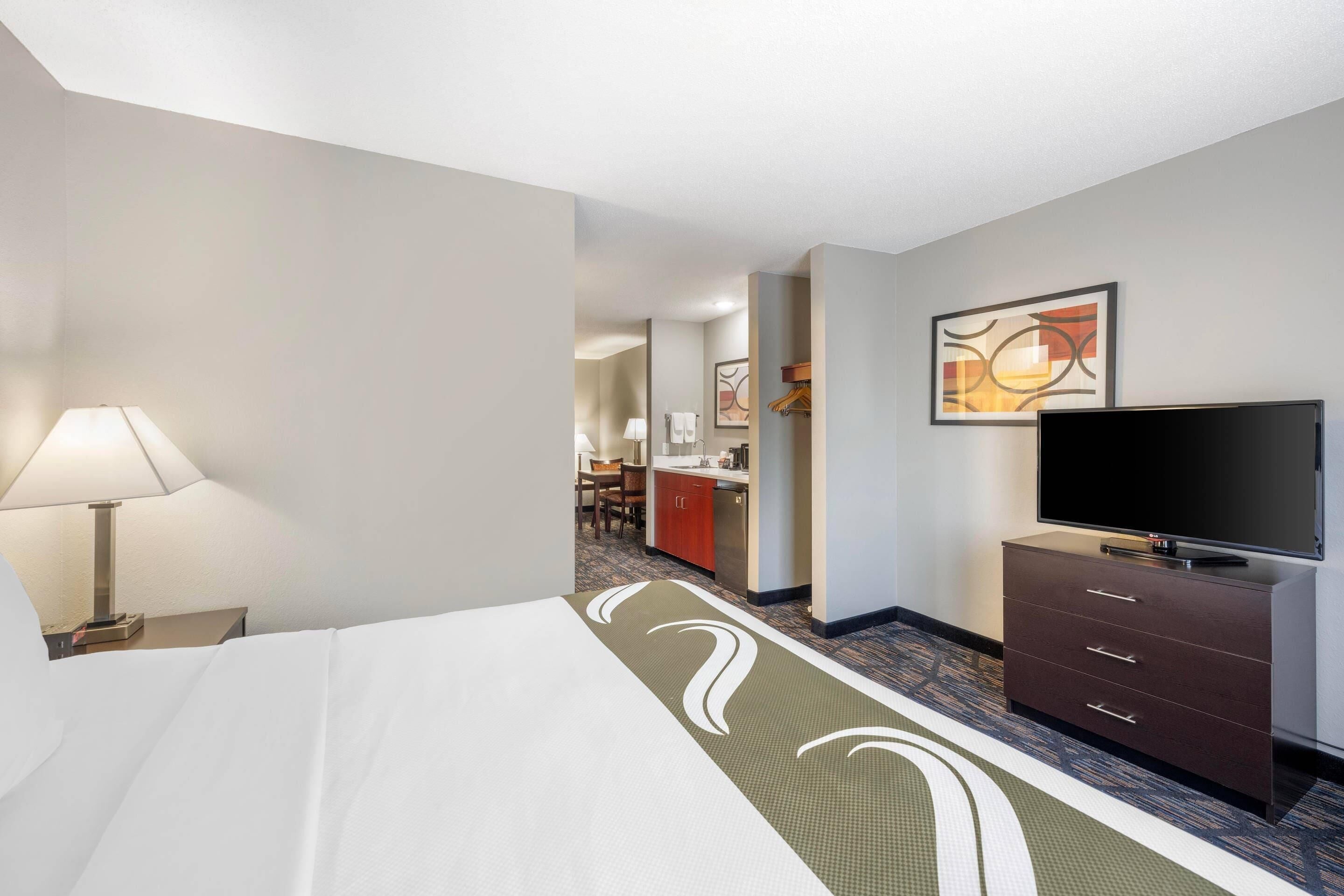 A hotel room with a bed, a TV on a dresser, a desk, and a kitchenette in the background.
