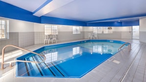 An indoor swimming pool with blue tiles, a metal ladder, and white chairs.