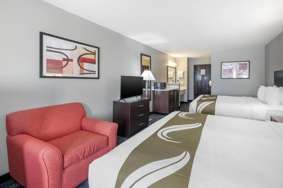 A hotel room with a red armchair, two beds, a TV, a desk with a lamp, and artwork on the walls.