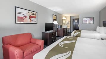 A hotel room with a red armchair, two beds, a TV, a desk with a lamp, and artwork on the walls.