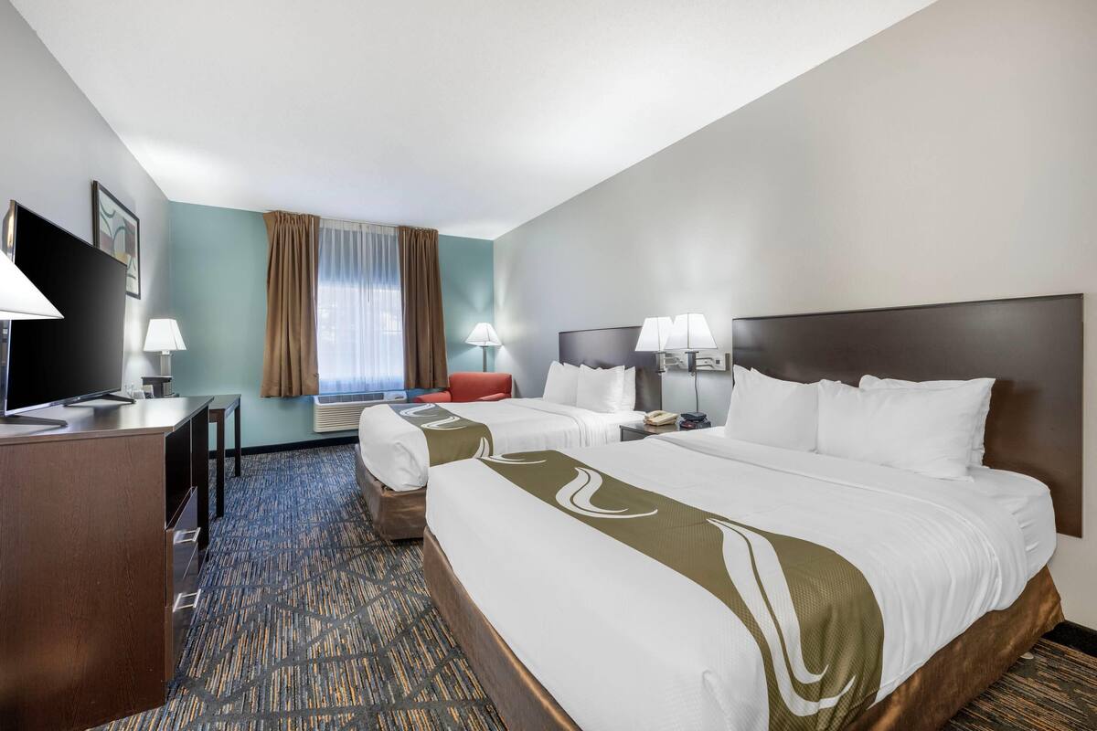 A hotel room with two beds, a desk, a TV, and a window with curtains.