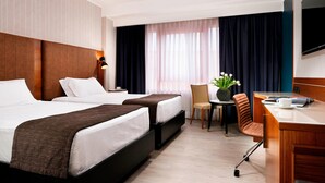 A hotel room with two beds, a desk, a chair, a TV, and a window with curtains. - Four Points by Sheraton Siena (Siena)