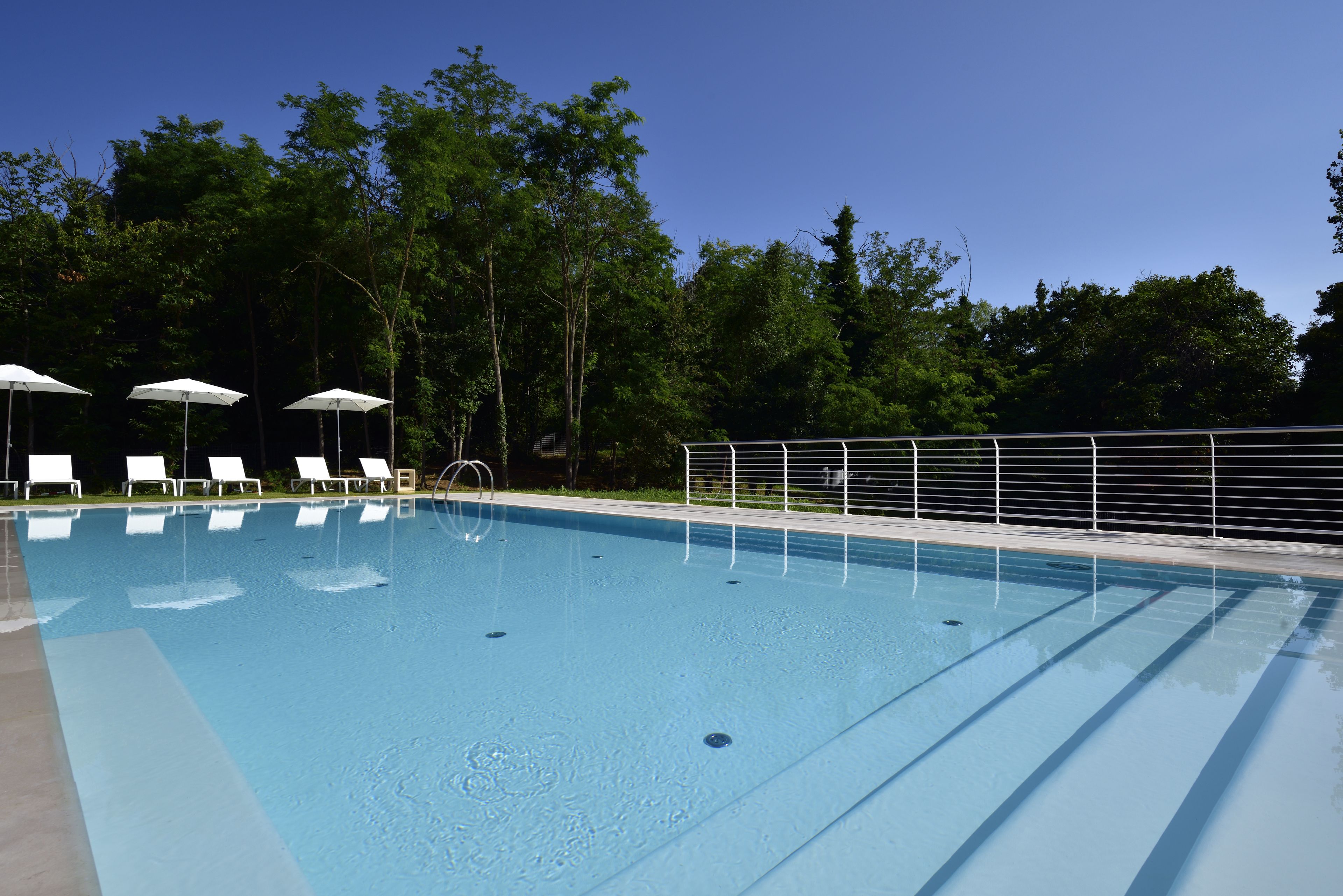 A clear blue swimming pool with white lounge chairs and umbrellas, surrounded by trees.