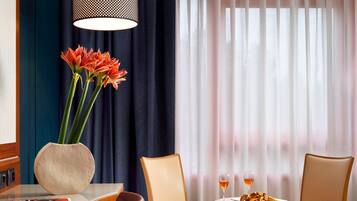Ein Hotelzimmer mit Esstisch, zwei Stühlen, einer Vase mit Blumen und einem Fenster mit Vorhängen.