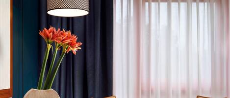A hotel room with a dining table, two chairs, a vase with flowers, and a window with curtains.