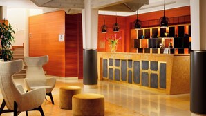 A reception area with a wooden reception desk, two chairs, a vase with flowers, and a large pillar. - Four Points by Sheraton Siena (Siena)