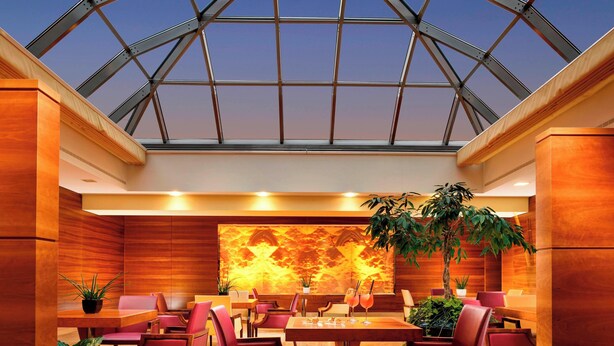 A modern hotel lobby with a high ceiling and a glass roof, featuring wooden paneling, comfortable seating, and a large wall mural. - Four Points by Sheraton Siena (Siena)