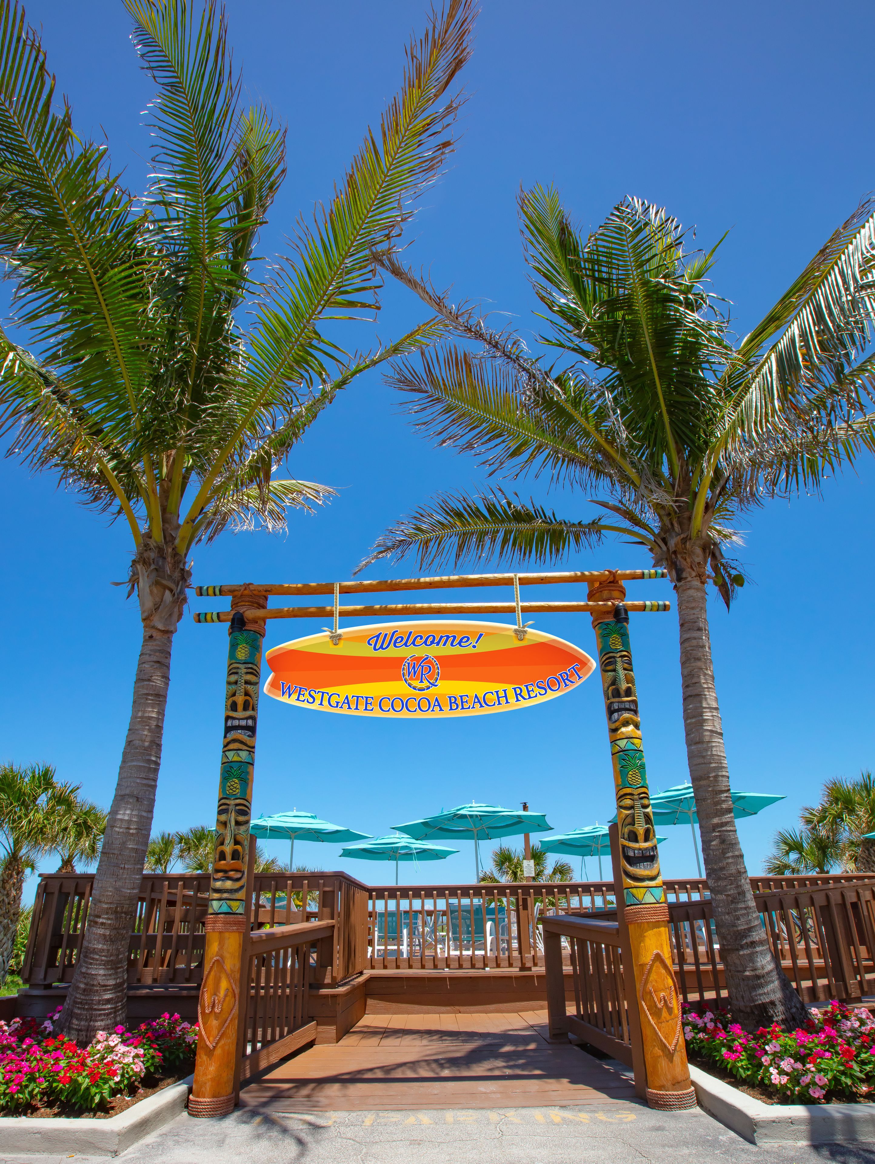 A tropical resort entrance with a welcoming sign, palm trees, and beach umbrellas.