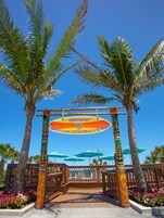 Entrada a un resort tropical con un letrero de bienvenida, palmeras y sombrillas de playa.
