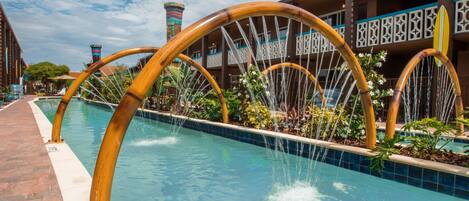 A pool with bamboo arches and water features, surrounded by a building with balconies and a tiled pool edge.