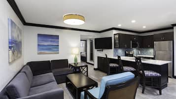A modern living room with a sofa, coffee table, and a dining area with chairs and a table. A kitchen with dark cabinets and stainless steel appliances is visible in the background.