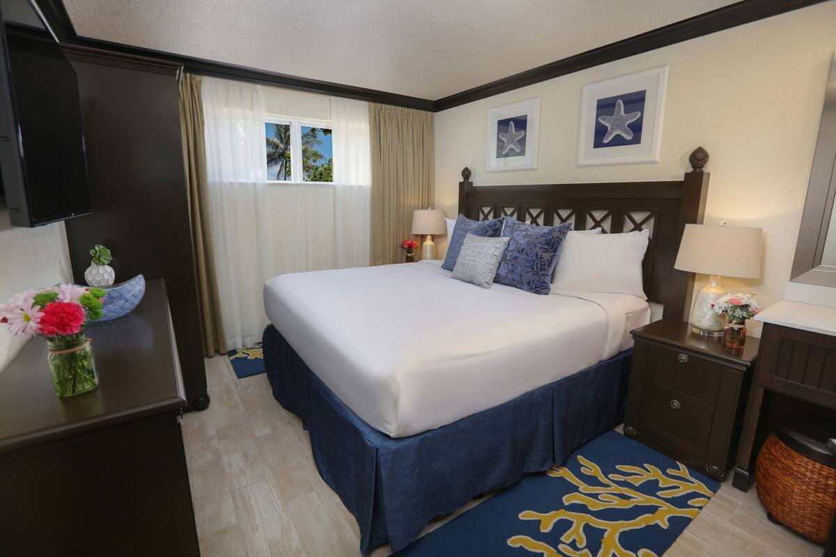 A hotel room with a large bed, a desk, a chair, a TV, a window with curtains, and a nightstand with a lamp and vase.