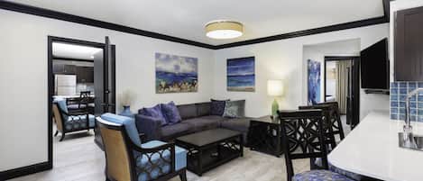 A modern living room with a sectional sofa, a coffee table, and a dining area with chairs. There is a kitchen visible in the background.