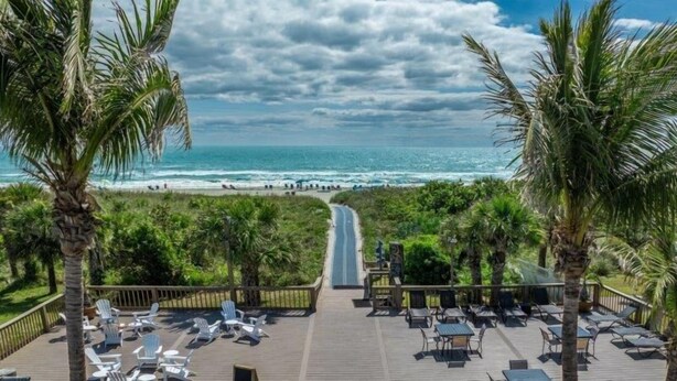 A deck with lounge chairs and tables overlooking a beach with palm trees and ocean waves. - Westgate Cocoa Beach Resort (Cocoa Beach)